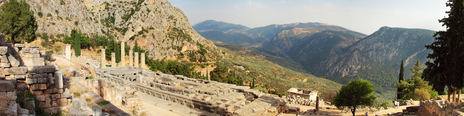 Ancient ruins with columns and stone structures, set against a backdrop of mountains and trees.