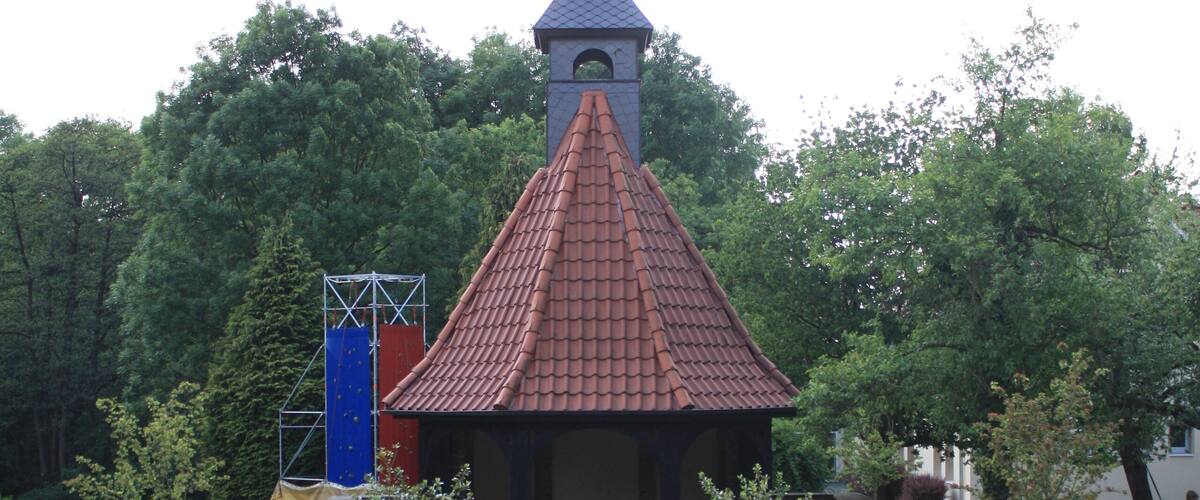 A pathway leading to a small building with a red-tiled roof and a cross on top, surrounded by greenery and benches.
