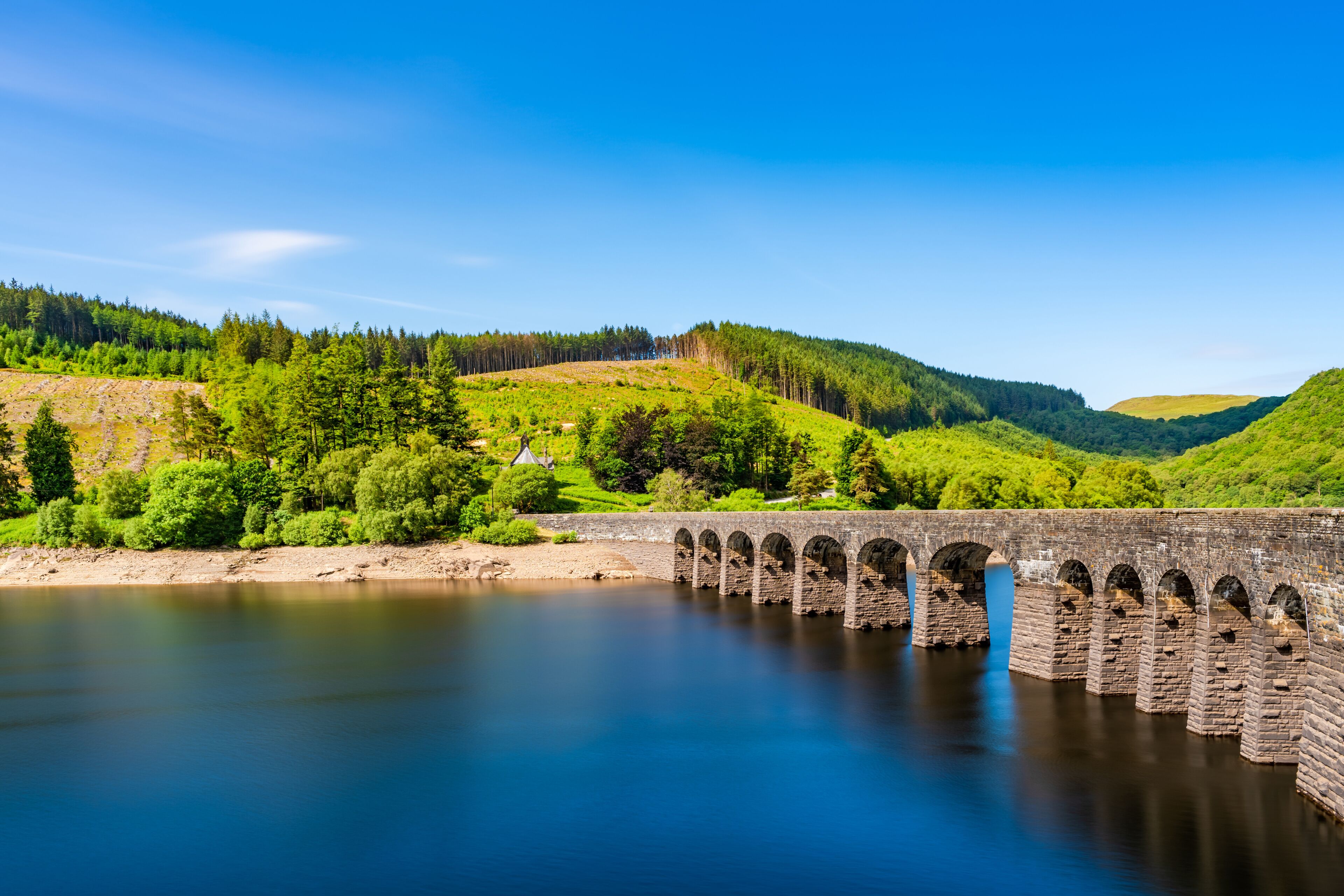 Un pont de pierre enjambe une rivière paisible, entouré de collines verdoyantes et de forêts denses.