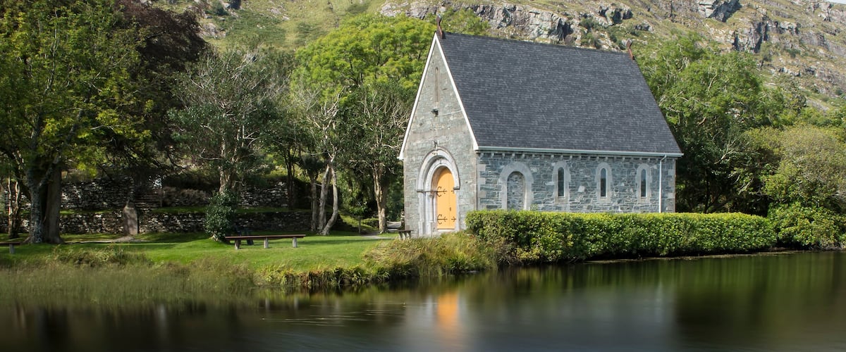 A stone church by a calm lake with red flowers in the foreground.