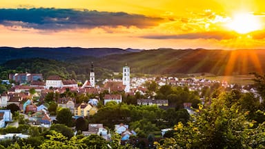 Une ville aux bâtiments aux couleurs vives, avec un clocher d’église imposant, et un coucher de soleil sur des collines ondoyantes.