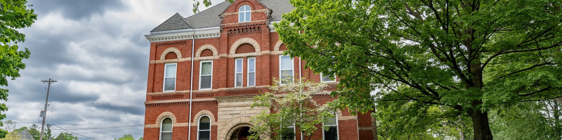 A historic red brick building with a clock tower, surrounded by lush green trees and a well-maintained lawn.