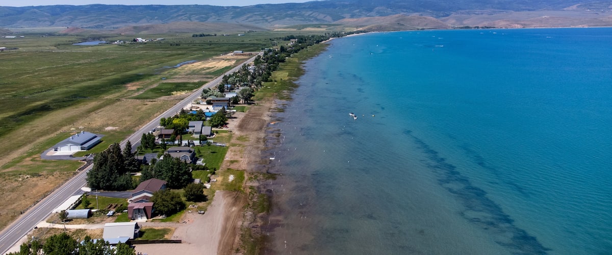 An aerial view of a coastal town with houses along the shoreline, a clear blue sea, and rolling hills in the background.