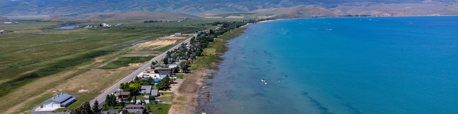 An aerial view of a coastal town with houses along the shoreline, a clear blue sea, and rolling hills in the background.