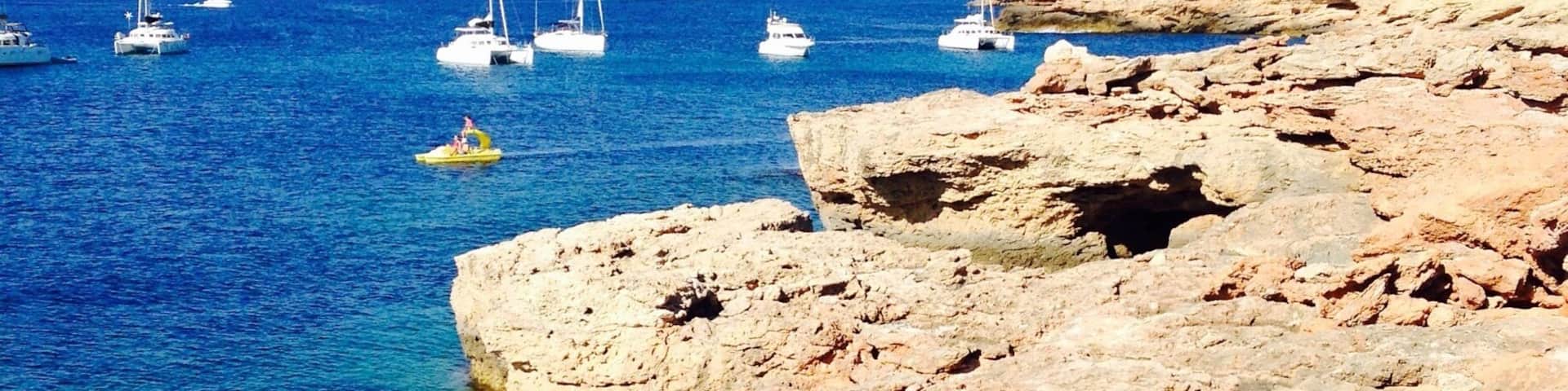A rocky coastline with clear blue waters and several sailboats anchored near the shore.