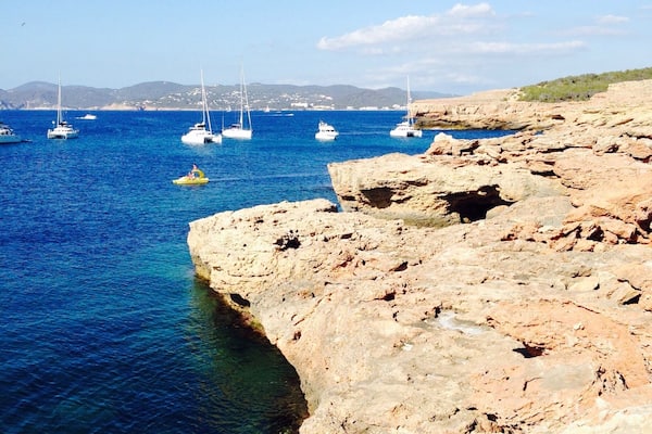 A rocky coastline with clear blue waters and several sailboats anchored near the shore.