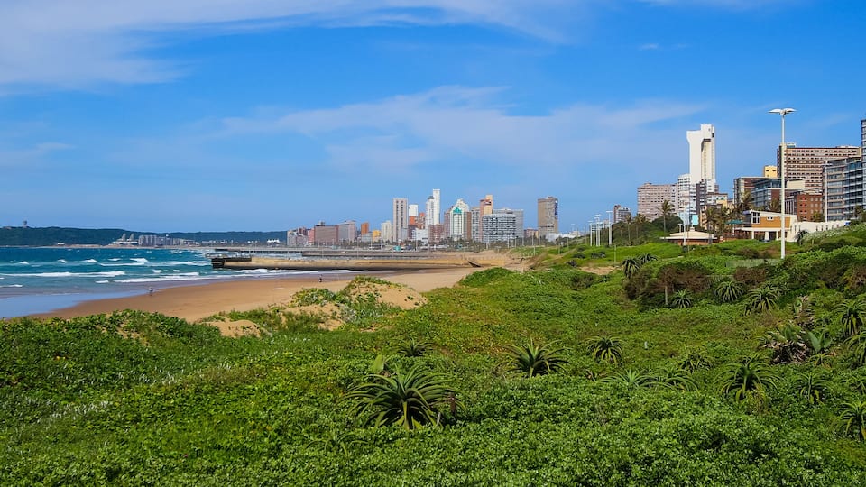 A coastal cityscape with a beach, dense greenery, and high-rise buildings.