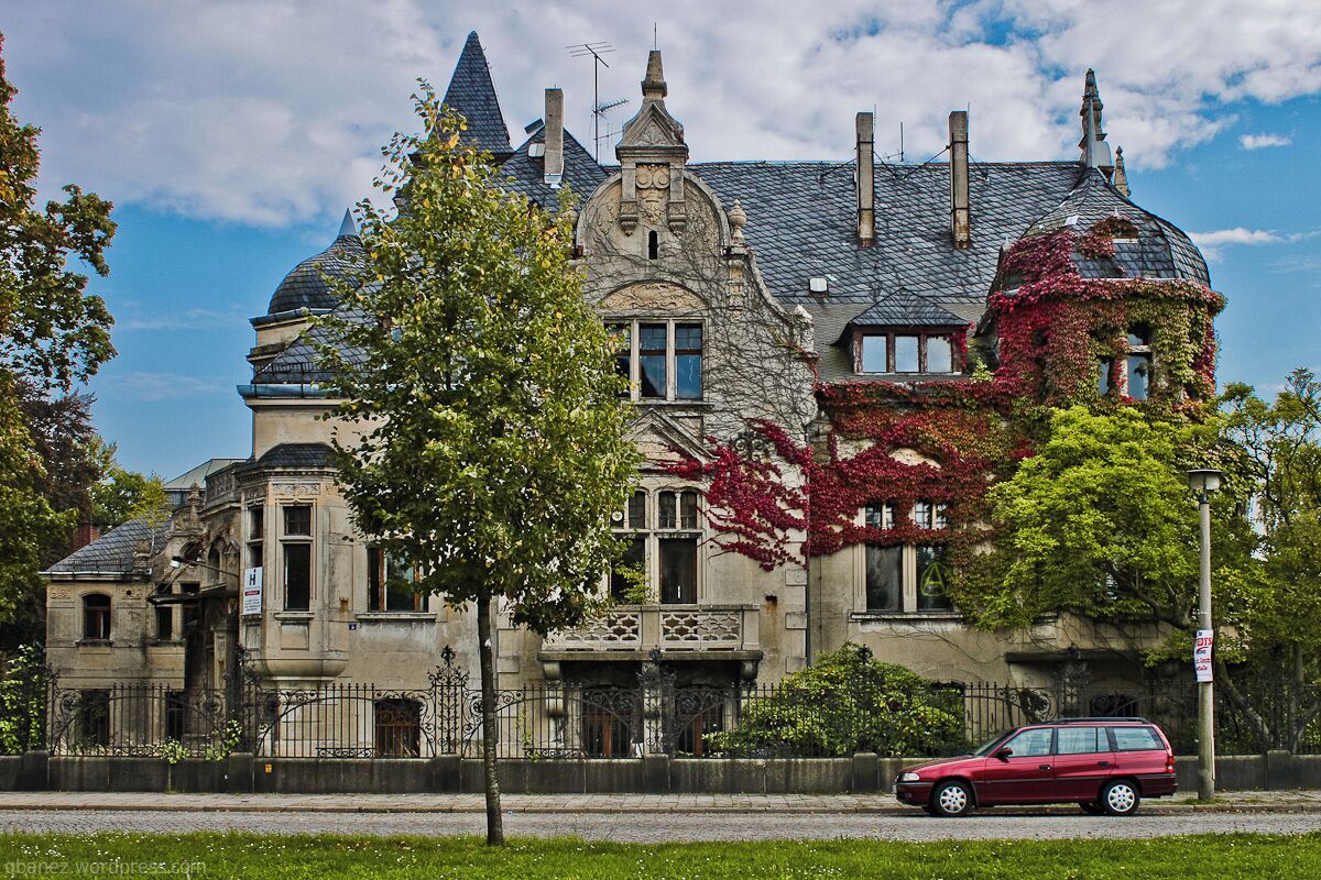 A historic stone building with a steeply pitched roof, ornate iron gates, and ivy-covered walls.