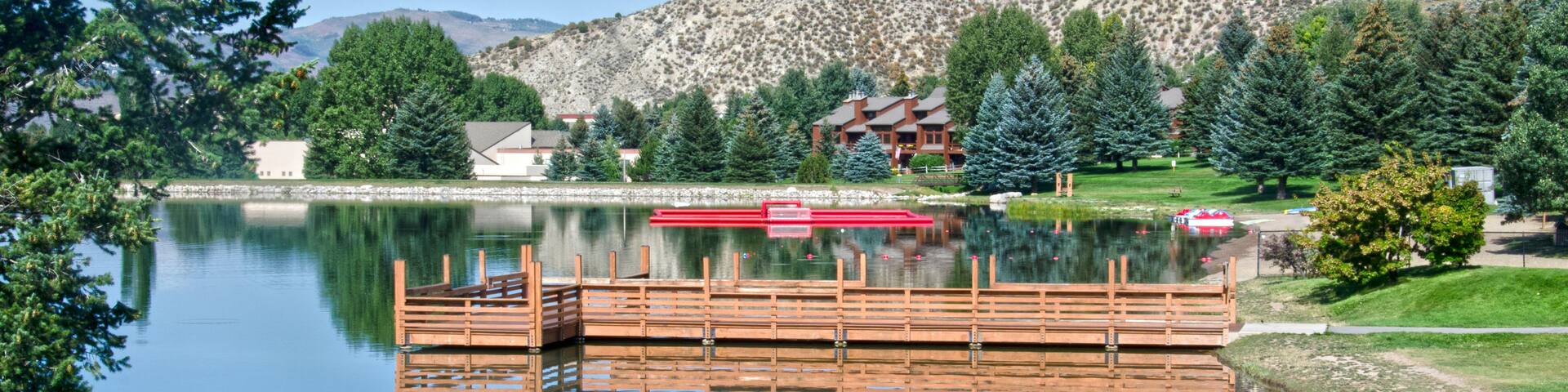 A calm lake with a wooden dock, surrounded by greenery and a mountain in the background.