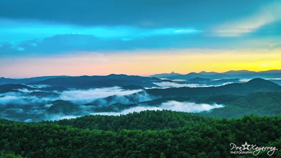A mountainous landscape with a forested valley and a sunrise or sunset sky.