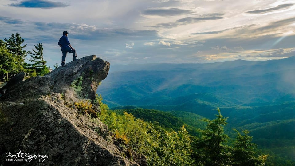 A person standing on a rocky outcrop overlooking a mountainous landscape with dense forest.