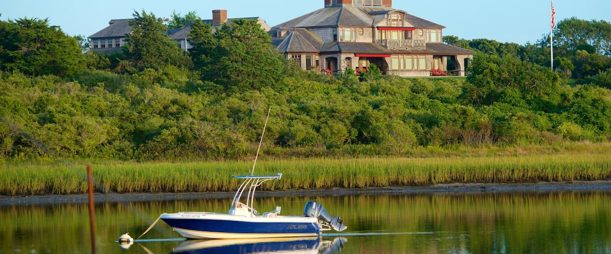A boat on a calm river, a large house with a red roof, and dense greenery in the background.