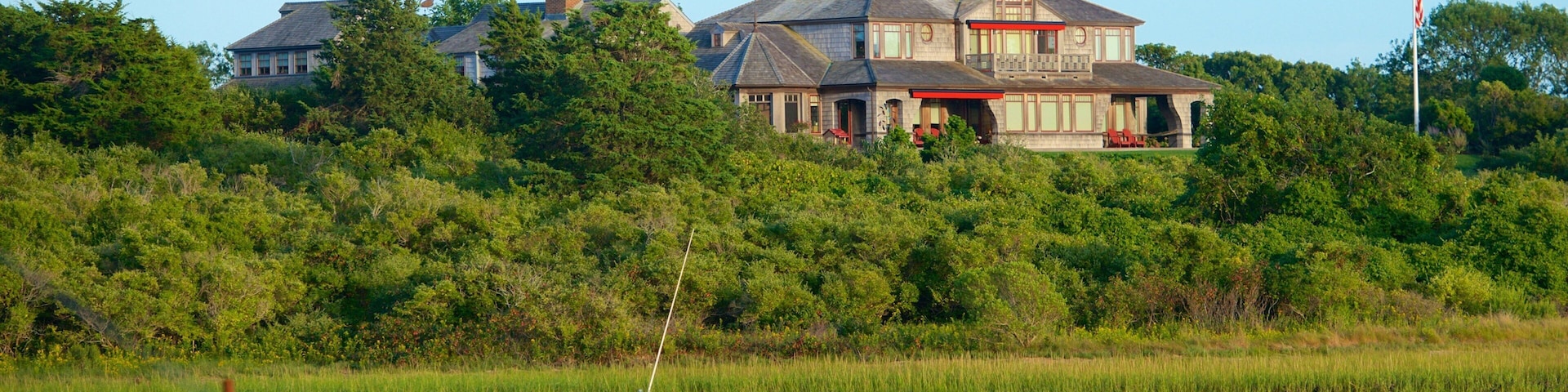A boat on a calm river, a large house with a red roof, and dense greenery in the background.