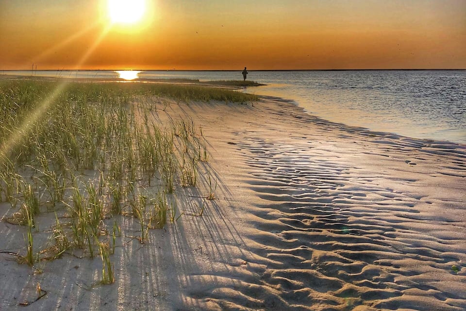 Una playa al atardecer, con el sol bajo en el horizonte proyectando largas sombras sobre la arena.