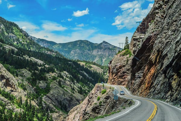 Une route de montagne sinueuse, avec une voiture, bordée de falaises rocheuses et de forêts denses.