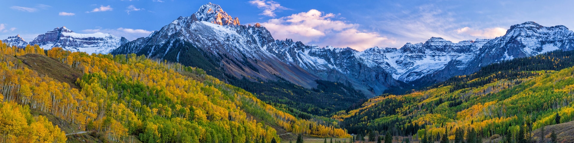 Uma cordilheira com picos nevados, um vale com árvores em tons de outono e um céu azul límpido.