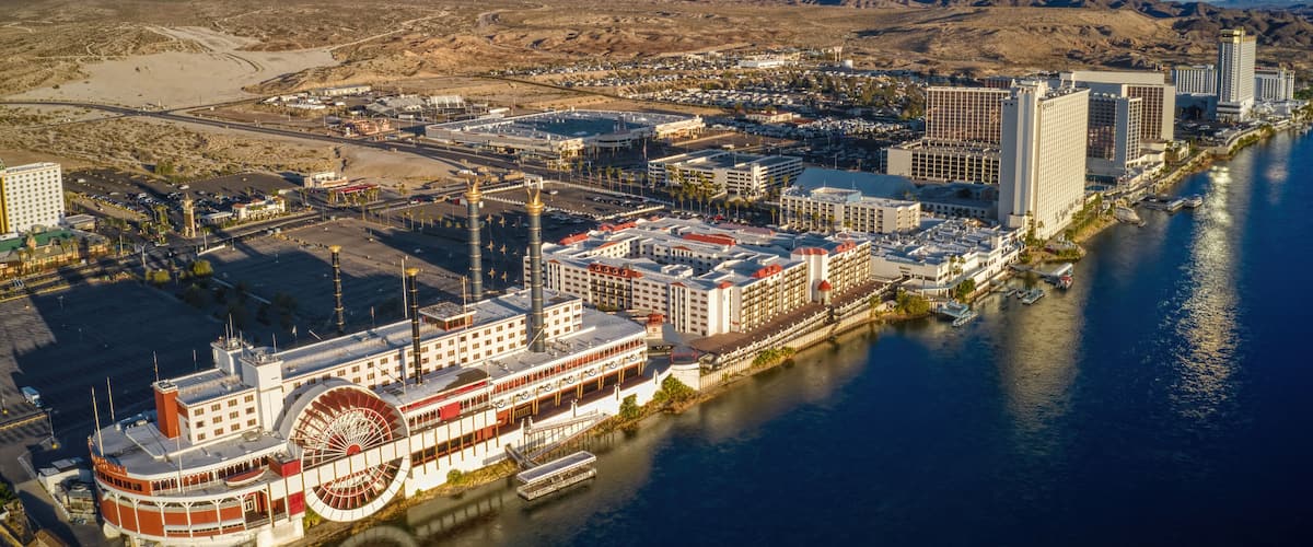 A riverboat docked by a river, surrounded by buildings and mountains in the background.