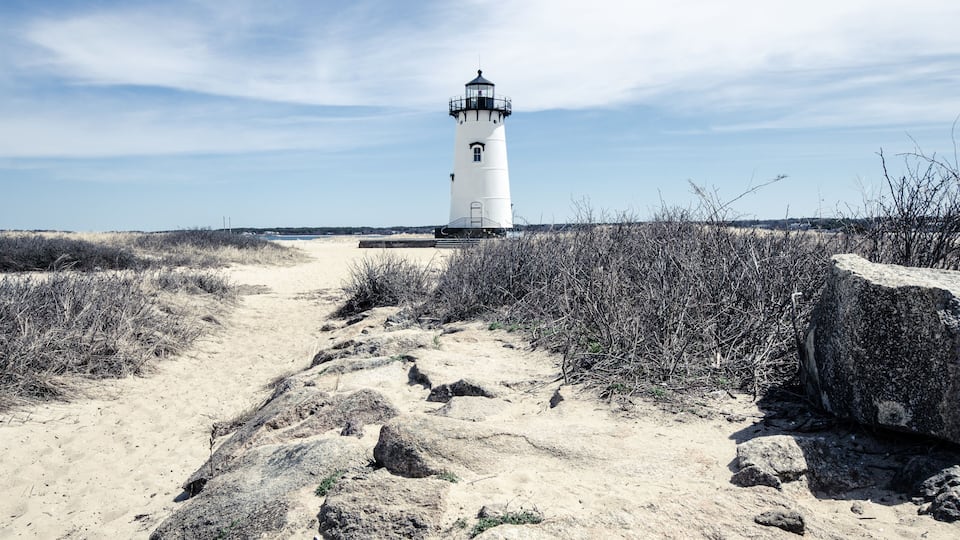 A white lighthouse on a sandy path with sparse vegetation and a clear sky.