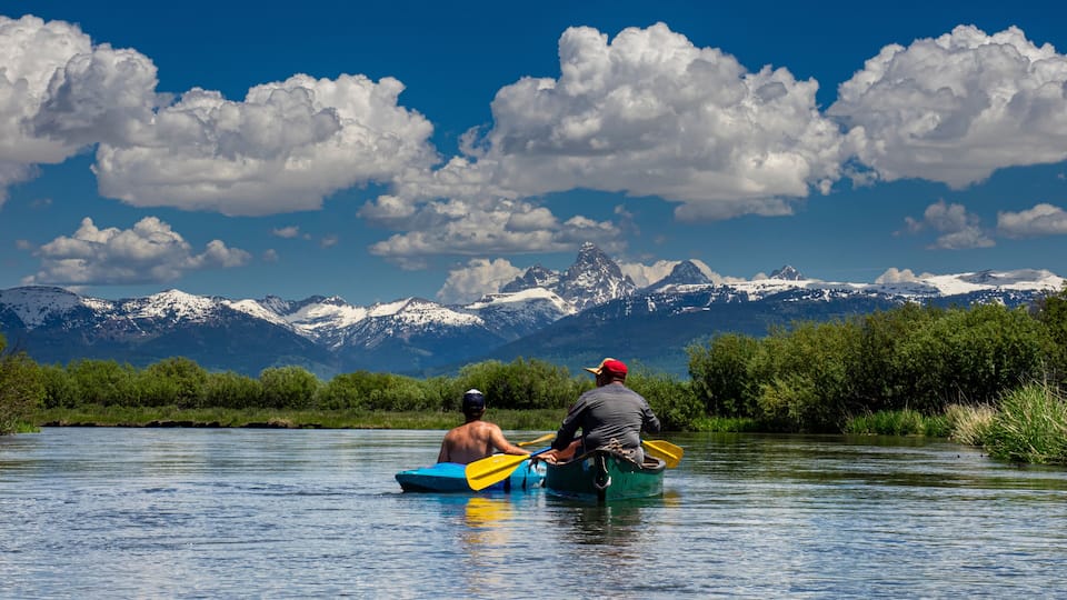 Two people kayaking on a calm river with a mountain range in the background.