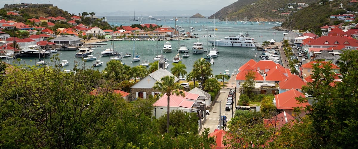 A coastal town with a marina filled with boats, surrounded by houses with red roofs, and a hilly landscape in the background.