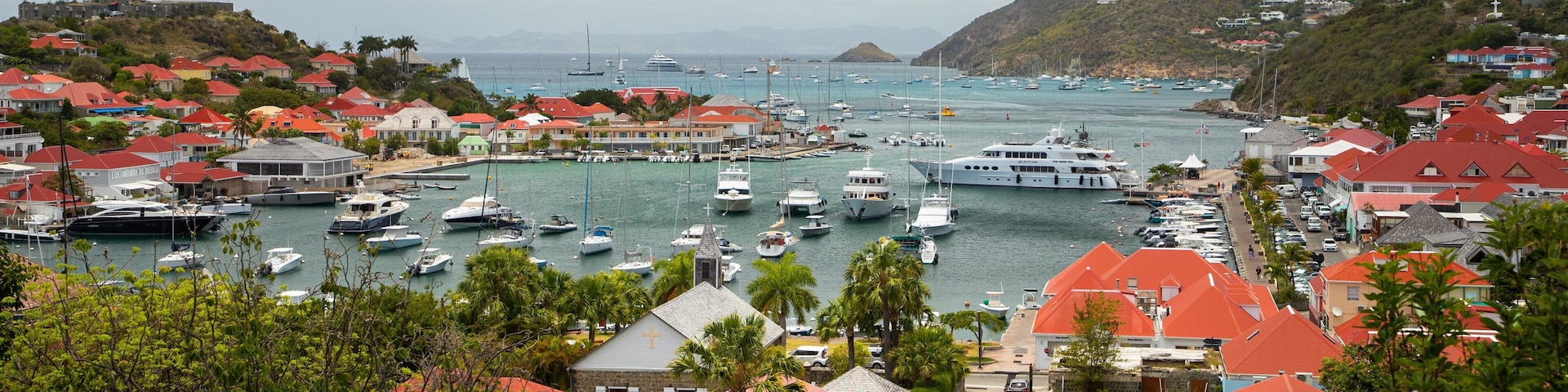 Un pueblo costero con un puerto deportivo lleno de barcos, rodeado de casas con tejados rojos, y un paisaje montañoso al fondo.