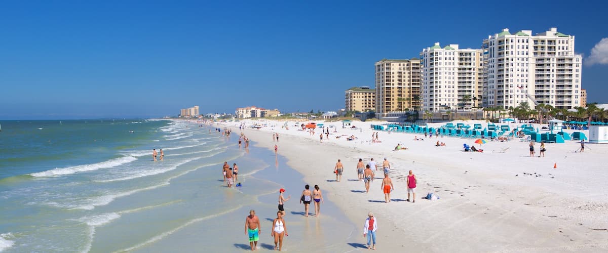 A crowded beach with people walking along the shore and buildings in the background.