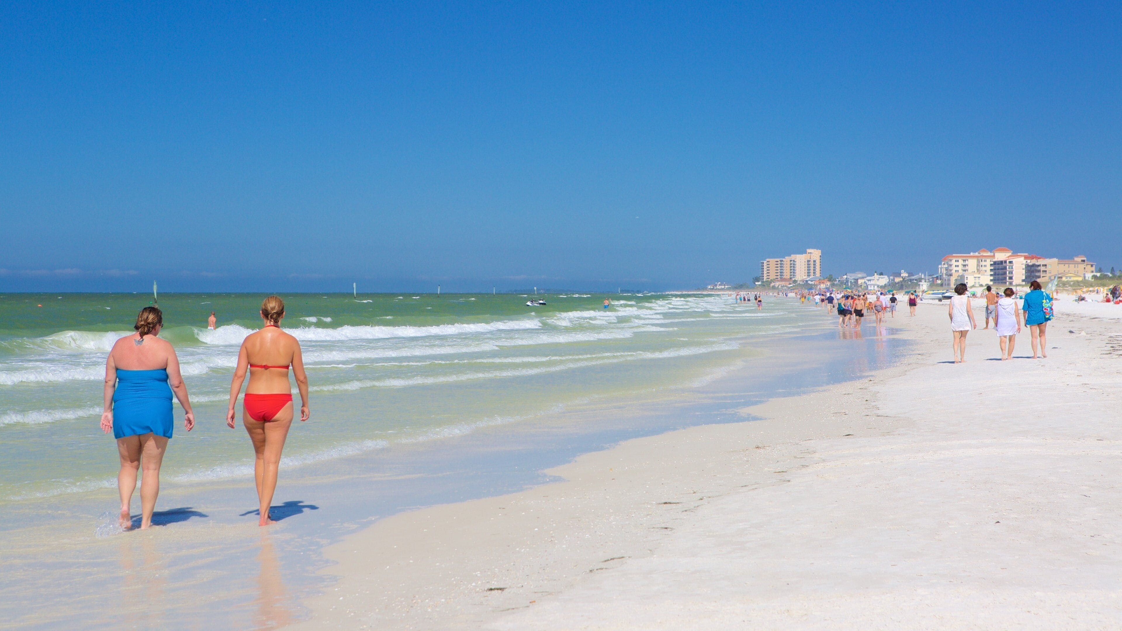 A beach with people walking along the shore, waves crashing onto the sand, and buildings in the background.