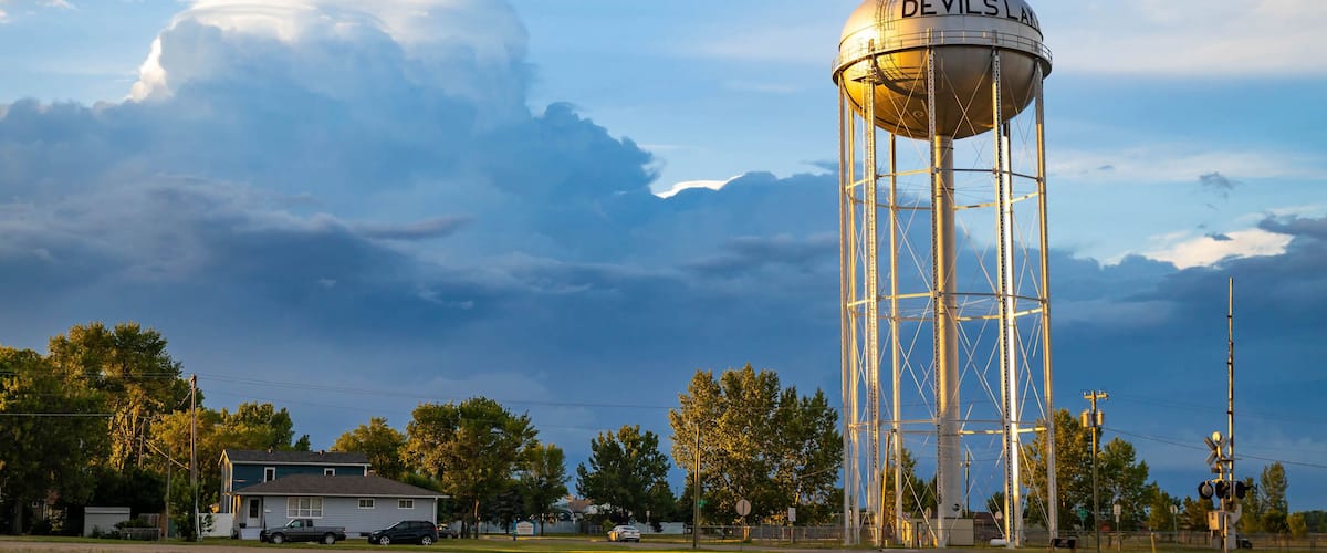 A water tower labeled "Devils Land" with a metal framework, a house, and a railroad track in the foreground.