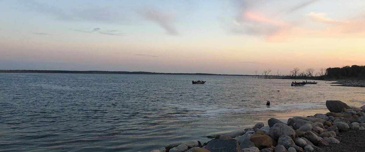 A calm river at sunset with boats and a rocky shoreline.