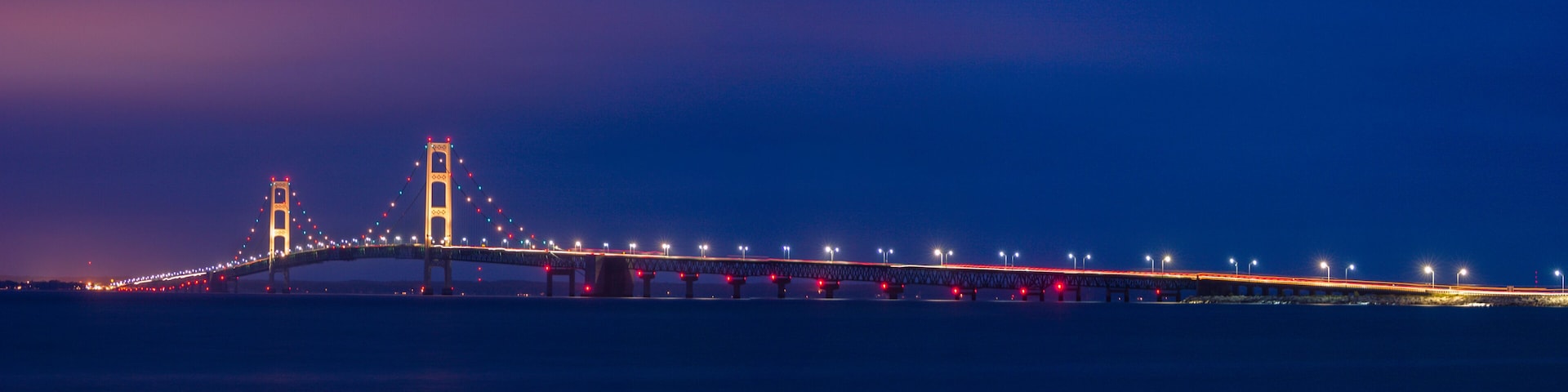 A long bridge with two towers, illuminated by lights against a twilight sky.