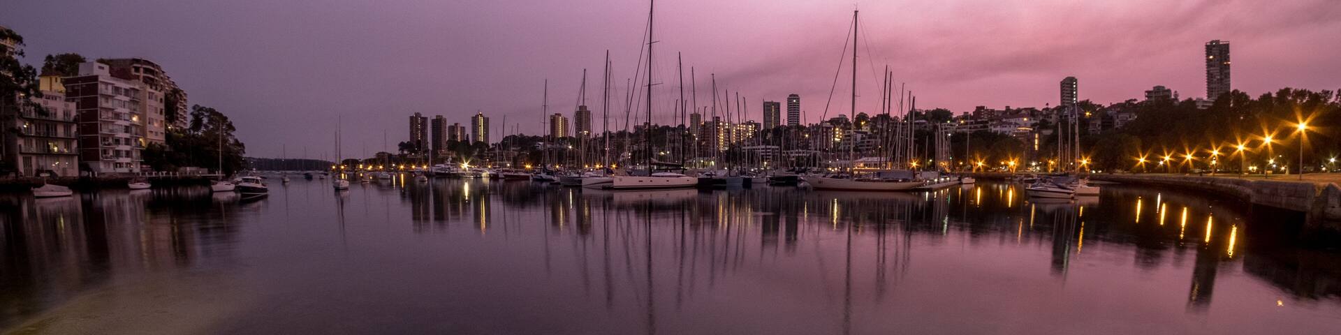 A marina at dusk with boats docked, city skyline in the background, and buildings along the waterfront.