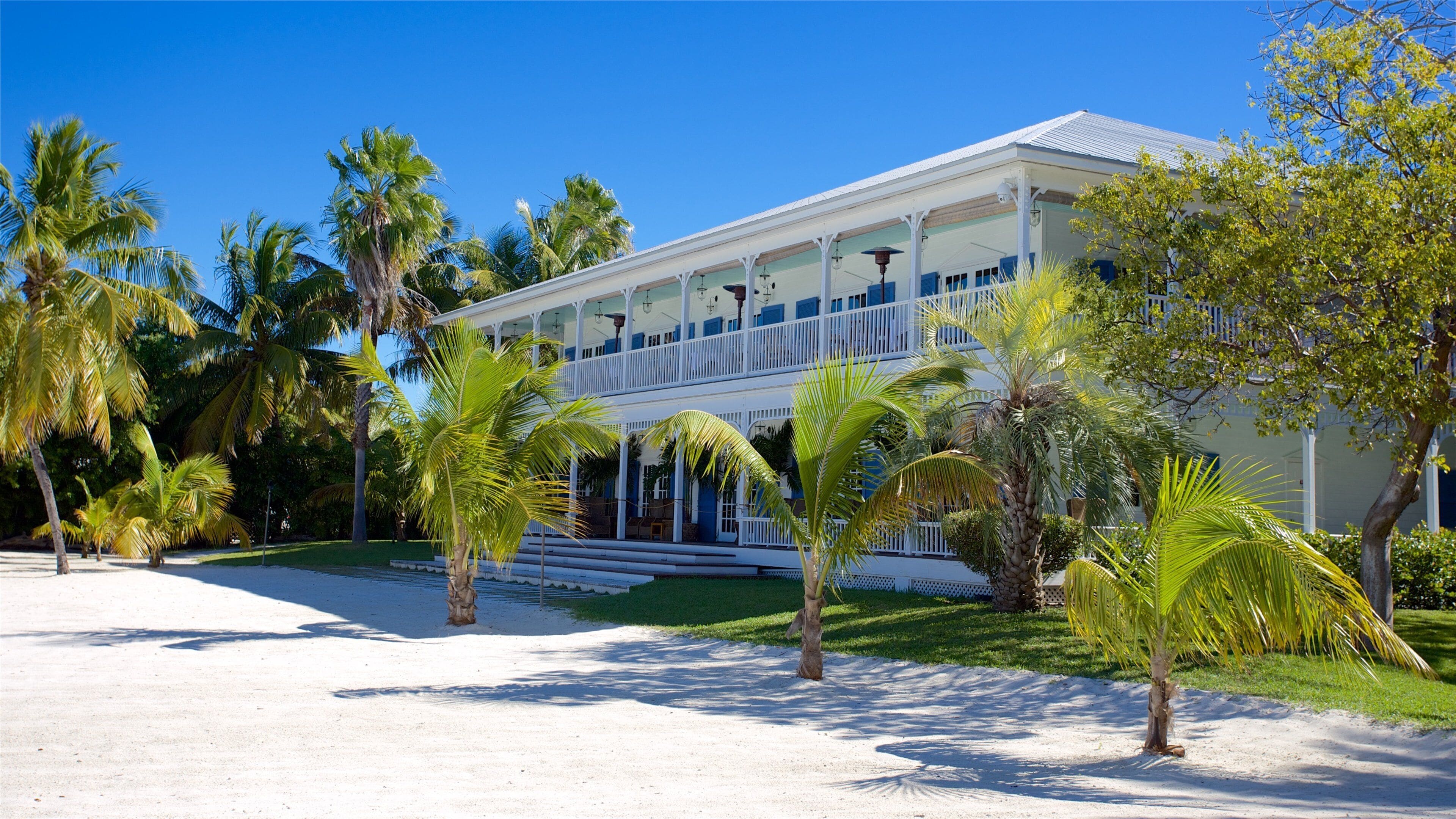 A white two-story house with blue accents, surrounded by palm trees and a sandy area.
