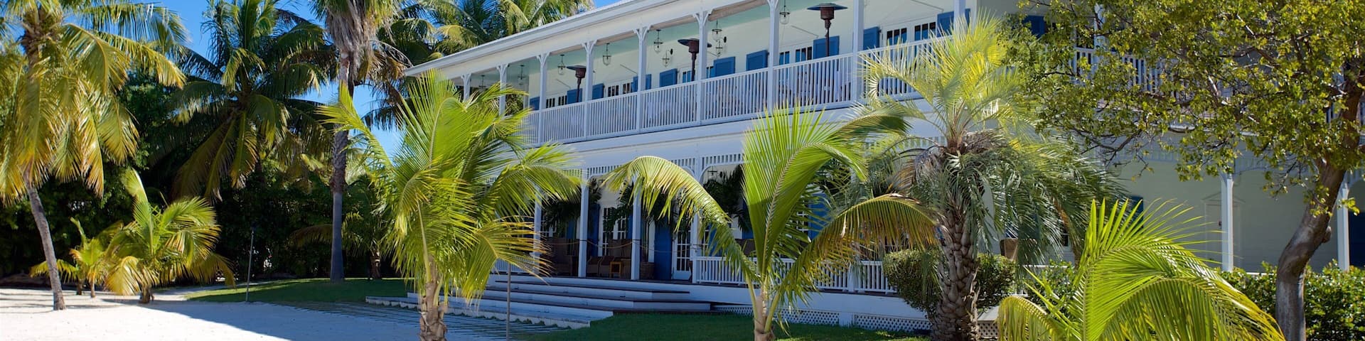 A white two-story house with blue accents, surrounded by palm trees and a sandy area.
