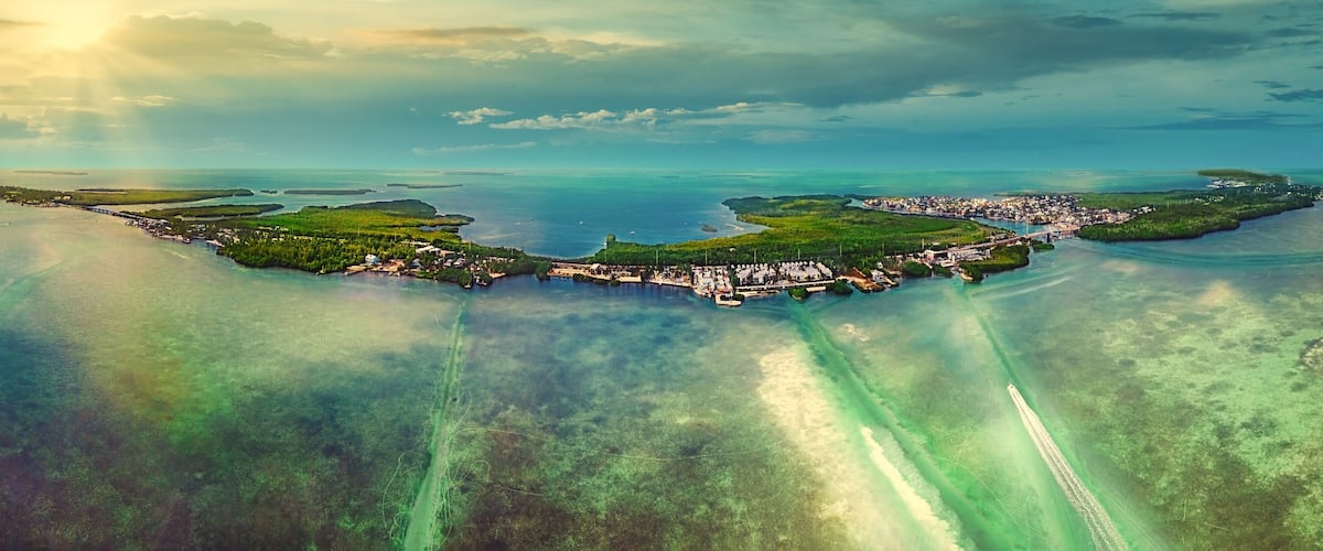 Vista aérea de un pueblo costero con un puerto deportivo, rodeado de aguas azules cristalinas y islas verdes.