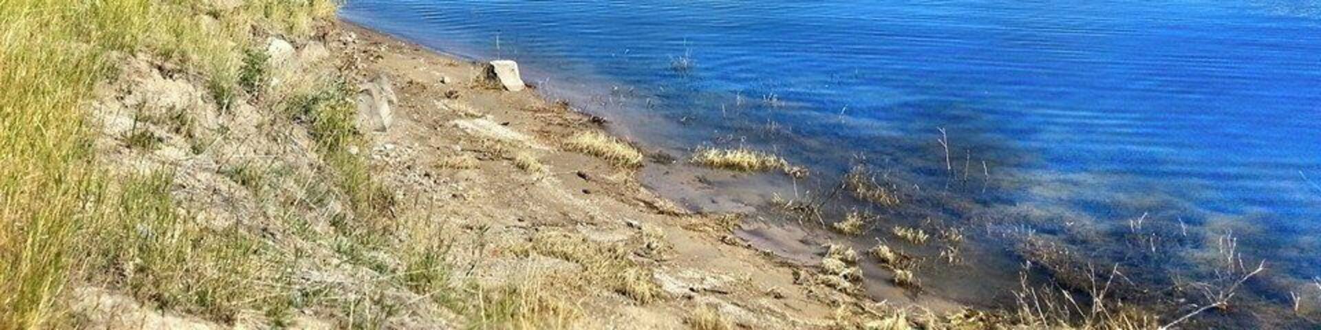 Un lago sereno con aguas azules cristalinas, rodeado de colinas cubiertas de hierba y montañas lejanas bajo un cielo despejado.