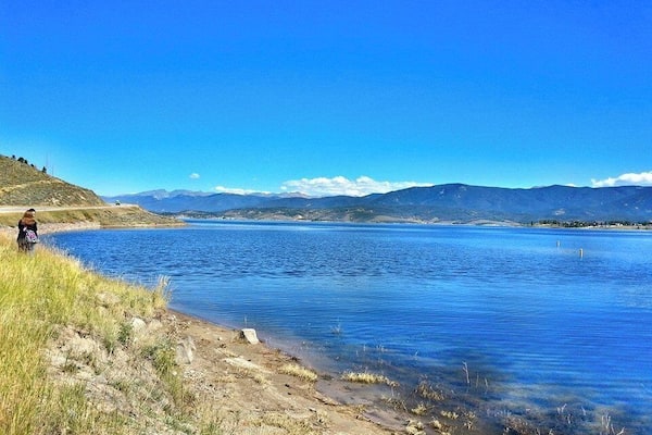 Un lac paisible aux eaux d’un bleu limpide, entouré de collines verdoyantes et bordé de montagnes lointaines, sous un ciel dégagé.