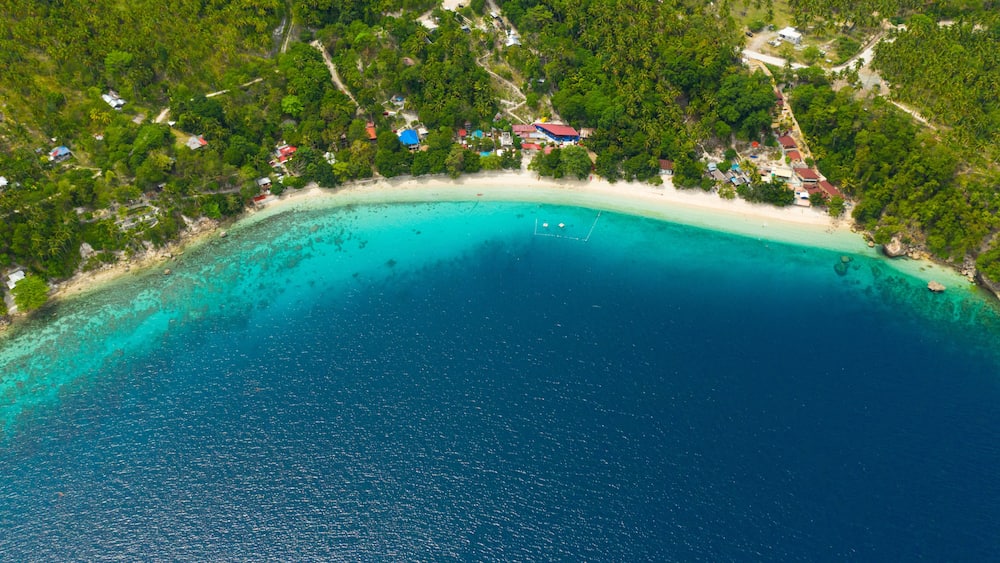 Une plage tropicale aux eaux turquoise et limpides, bordée d’une végétation luxuriante et ponctuée de quelques bâtiments dispersés.