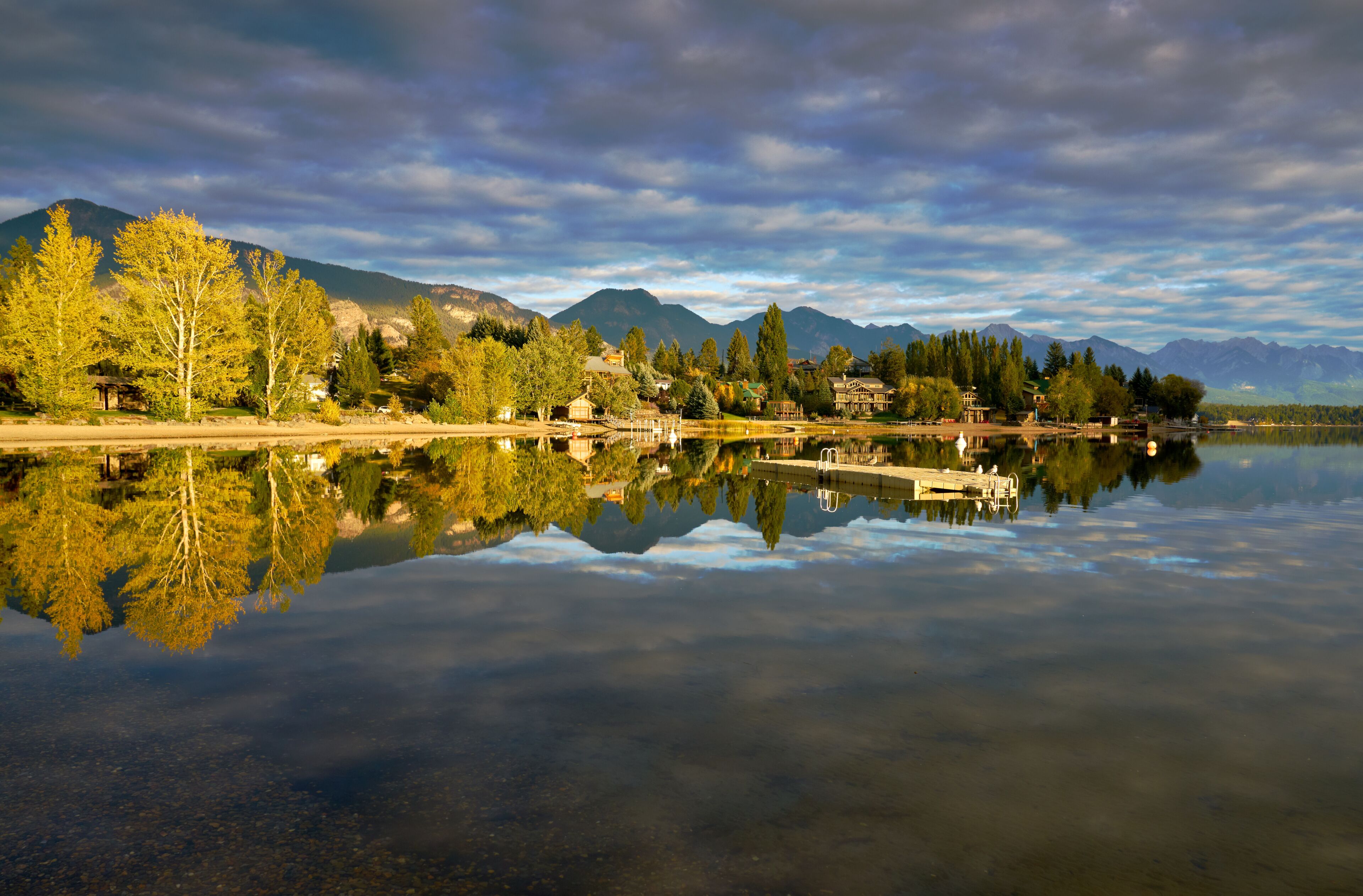 A calm lake with a reflection of trees and mountains, a dock, and houses along the shore.