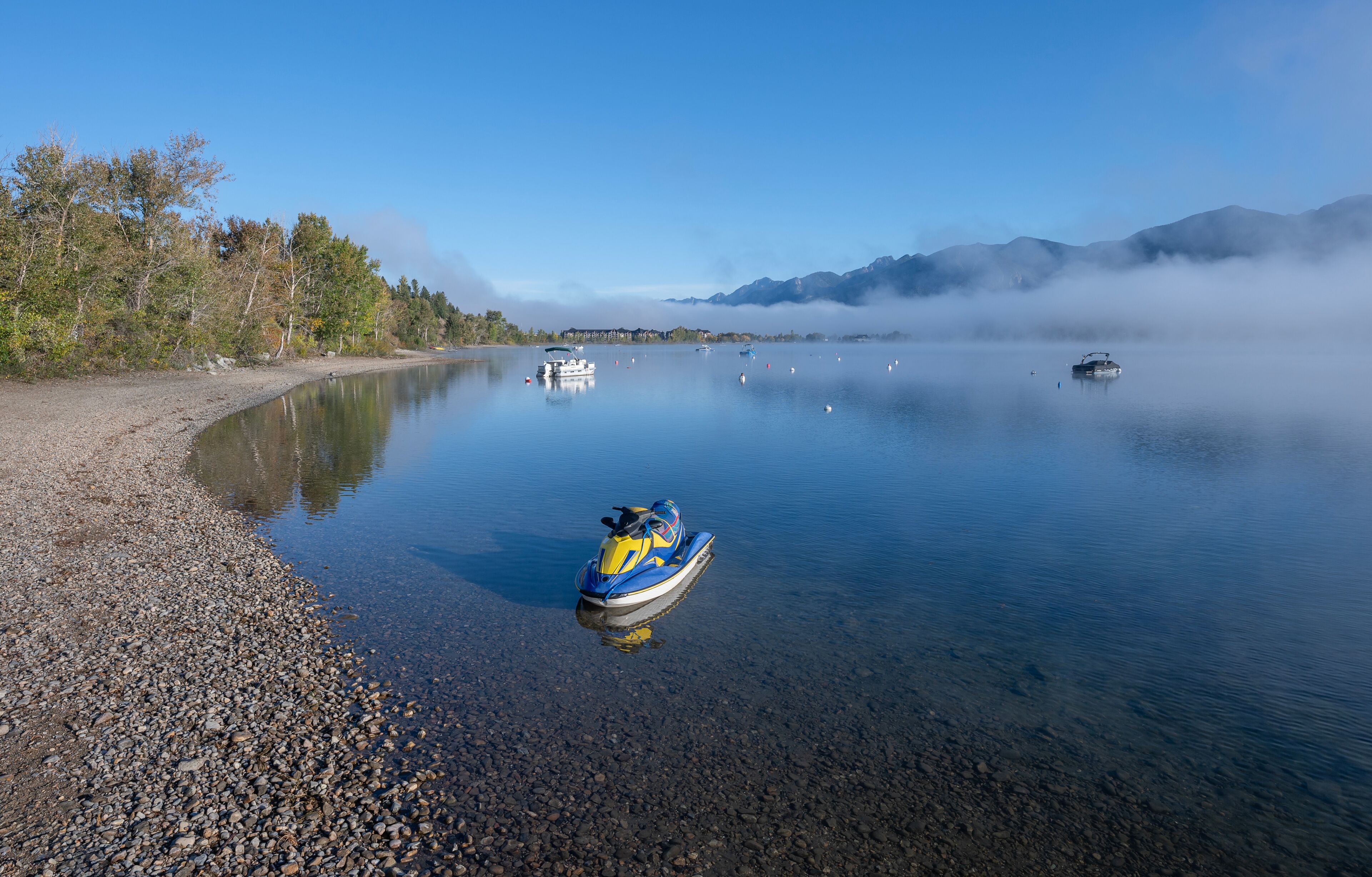 A calm lake with a jet ski on the shore, boats on the water, and mountains in the background.