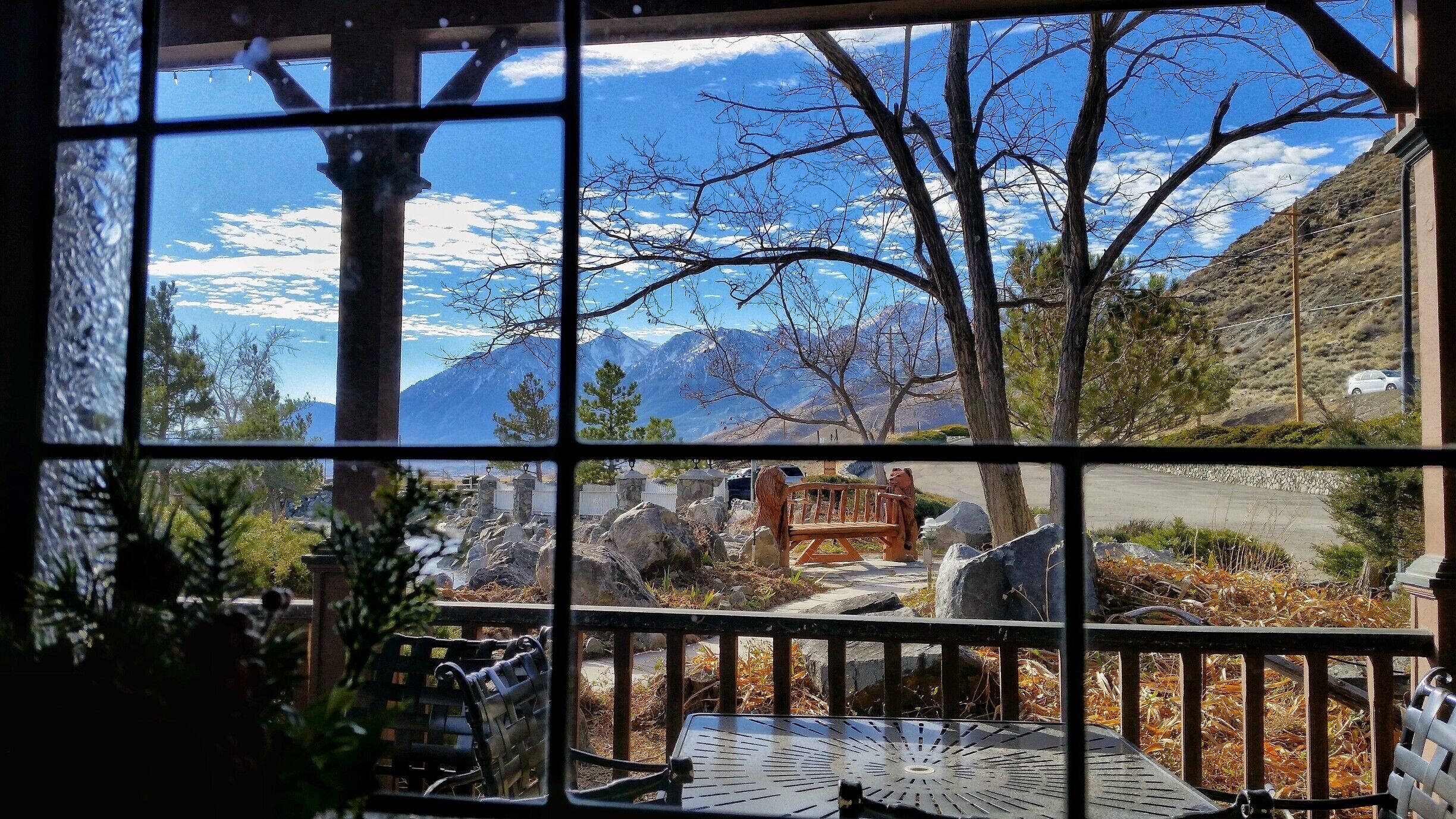 A scenic view through a window with mountains in the background, a wooden bench, and a stone pathway.