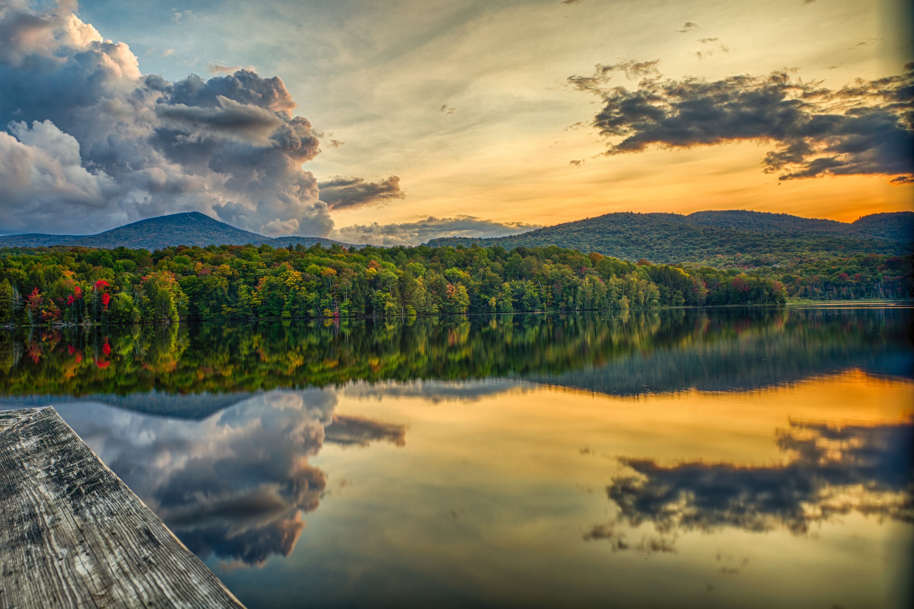 Un lago tranquilo que refleja árboles y montañas, con un muelle de madera en primer plano.