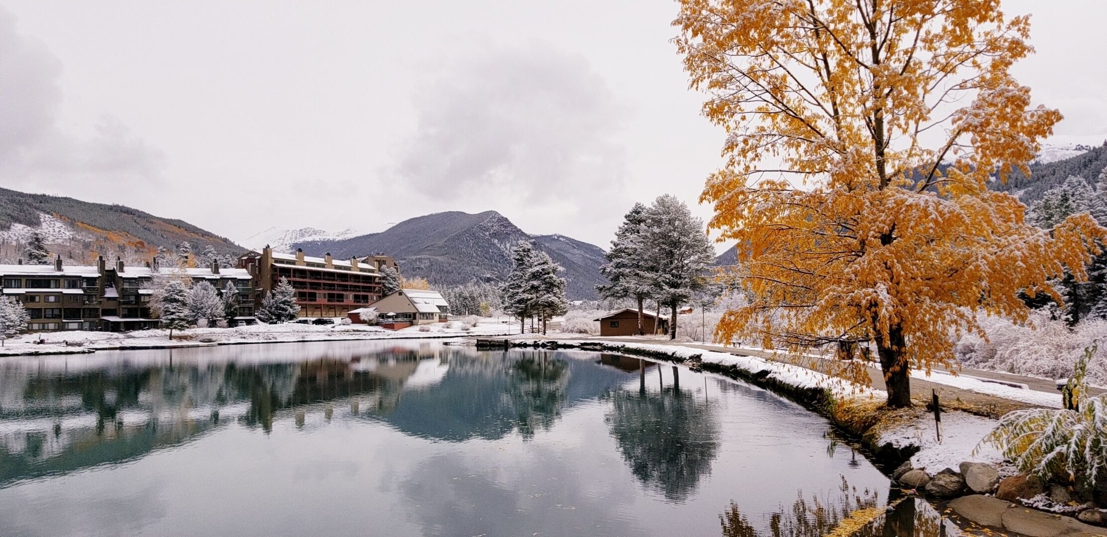A winter scene with a snow-covered lake, buildings, and mountains in the background.