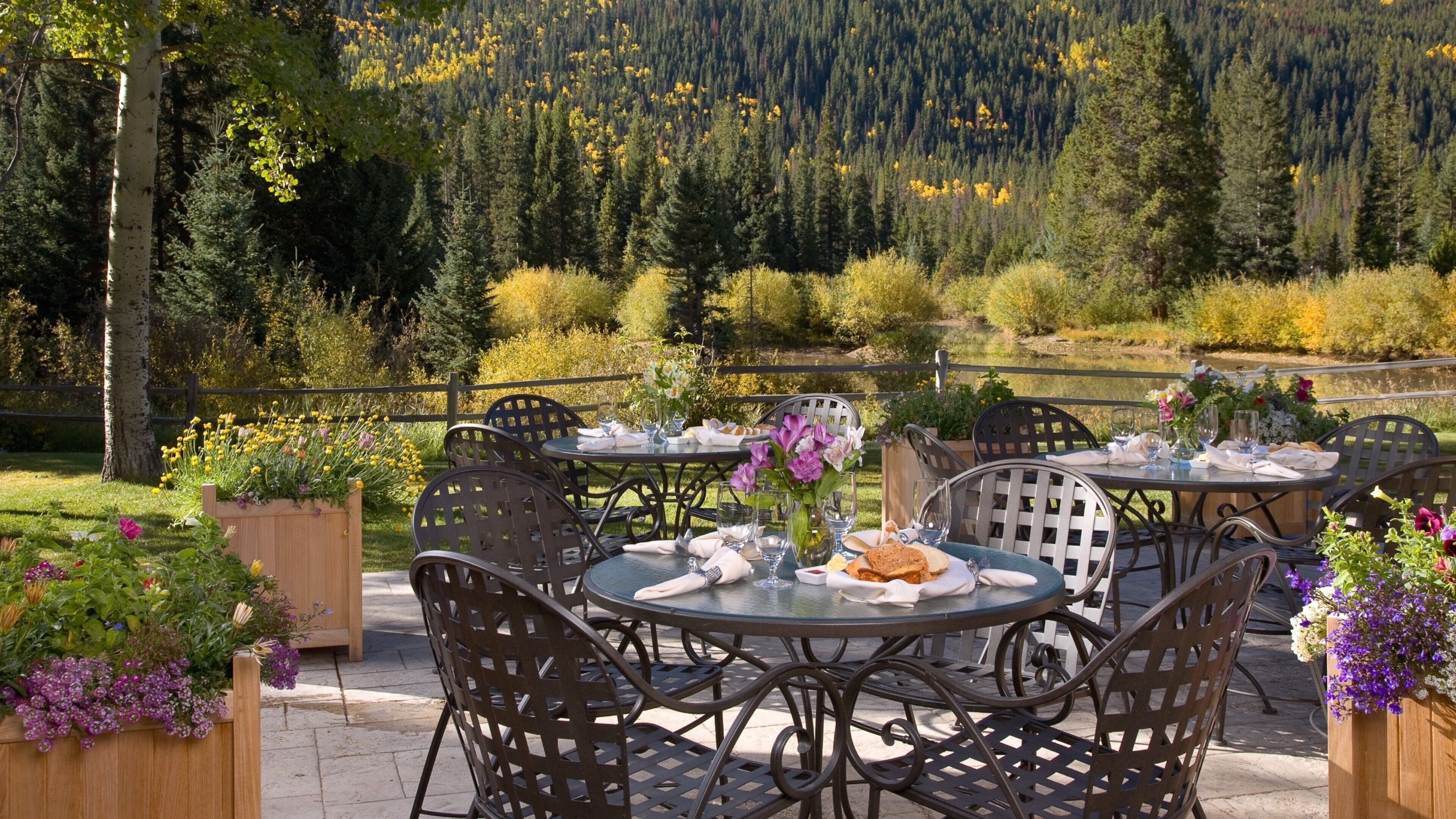 An outdoor dining area with round tables and chairs, surrounded by flower planters and a scenic view of a forested hillside.
