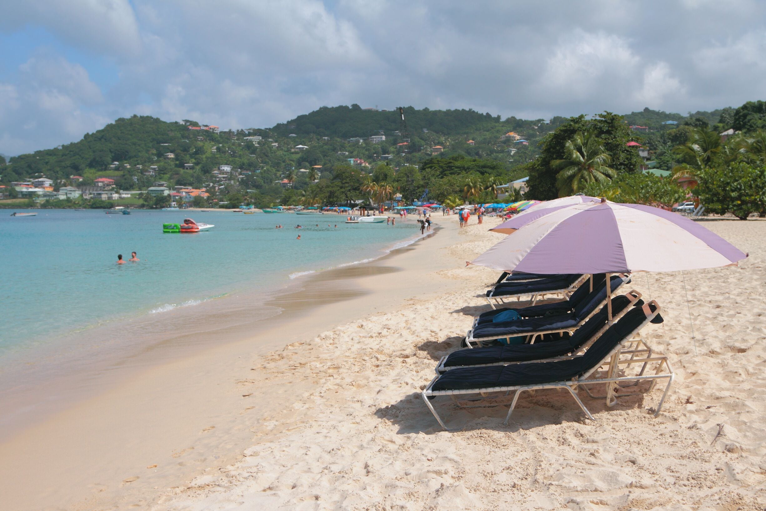 Uma praia com cadeiras de praia e um guarda-sol roxo, águas calmas e uma colina com casas.