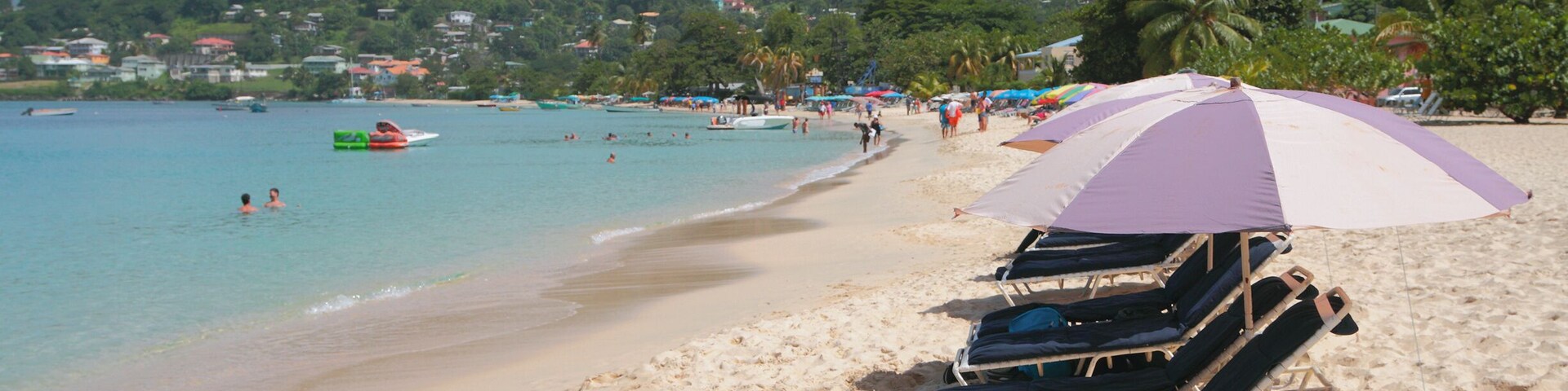 Uma praia com cadeiras de praia e um guarda-sol roxo, águas calmas e uma colina com casas.