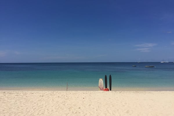 Une plage avec deux planches de surf appuyées contre un poteau, un ciel bleu dégagé et une mer calme avec des bateaux à l’horizon.