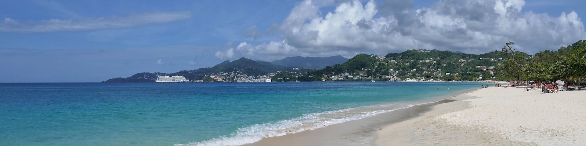 A beach with clear turquoise water, a sandy shore, and a backdrop of green hills and a distant mountain range under a partly cloudy sky.