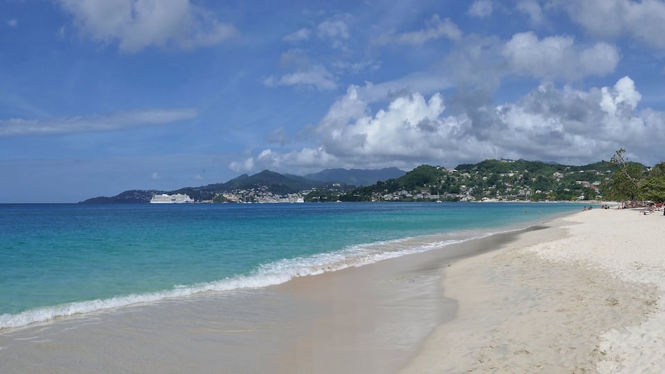 Una playa con aguas turquesas cristalinas, una orilla arenosa y un telón de fondo de colinas verdes y una cadena montañosa distante bajo un cielo parcialmente nublado.