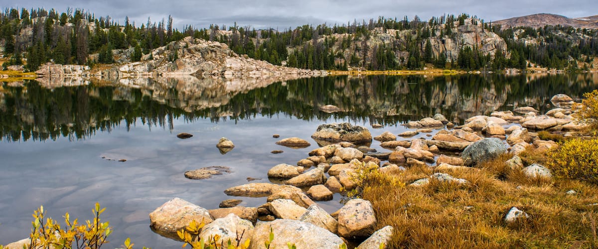 A calm lake with clear reflections, surrounded by rocky terrain and dense forest.