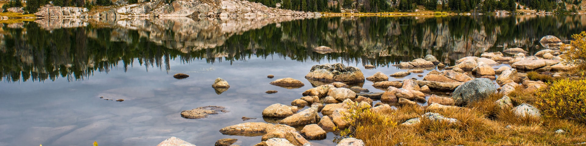 A calm lake with clear reflections, surrounded by rocky terrain and dense forest.